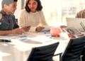 Three colleagues around a conference table review documents with a laptop and a pink piggy bank on the table in a bright office.