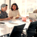 Three colleagues around a conference table review documents with a laptop and a pink piggy bank on the table in a bright office.